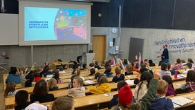 Ein Hörsaal voller Kinder, einige heben die Hände, der Dozent mit Mikrofon in der Hand läuft nach vorne zur Präsentation.