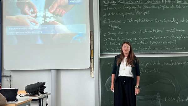Eine Frau vor einer beschriebenen Tafel, links von ihr eine Leinwand mit Präsentation. 