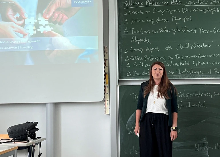 Eine Frau vor einer beschriebenen Tafel, links von ihr eine Leinwand mit Präsentation. 