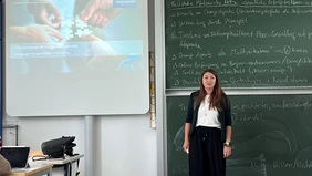 Eine Frau vor einer beschriebenen Tafel, links von ihr eine Leinwand mit Präsentation. 