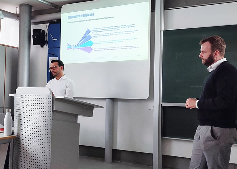 Zwei männliche Personen vor einem Whiteboard, auf dem eine Präsentation gezeigt wird.