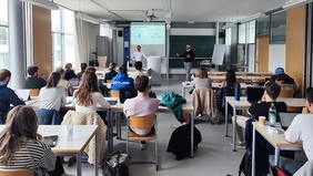 Studierende in einem Seminarraum, vorne zwei Referierende, von hinten aufgenommen.
