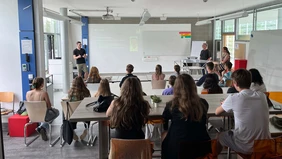 Im Hörsaal: von hinten sieht man Schülerinnen und Schüler, die dem Vortrag eines Mannes vorne an der Tafel zuhören.