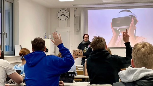 Eine Situation in einem Hörsaal – man sieht von hinten Studierende, die sich melden, vorne eine Frau an einem Laptop, hinter ihr eine Präsentation, die eine Person mit VR-Brille zeigt. 