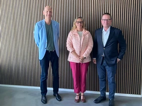 Gruppenbild bestehend aus drei Personen vor einer mit Holz verkleideten Wand