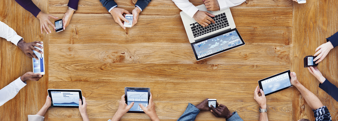 Blick auf einen Holzschreibtisch von oben, an dem Hände, Notebooks, Tablets und Mobiltelefone zu sehen sind