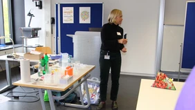 Die Professorin steht an einem Tisch voller Laborutensilien und Haushaltsmittel. Auf einem anderen Tisch sieht man eine Tüte Erdbeeren.