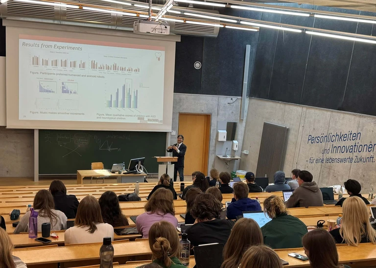 View from the back of the lecture hall, a presentation on the screen at the front, a speaker in a suit at the lectern, the students can be seen from behind  