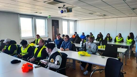 Man sieht eine Gruppe Studierender in einem Besprechungszimmer sitzen, teilweise tragen sie gelbe Warnwesten. 