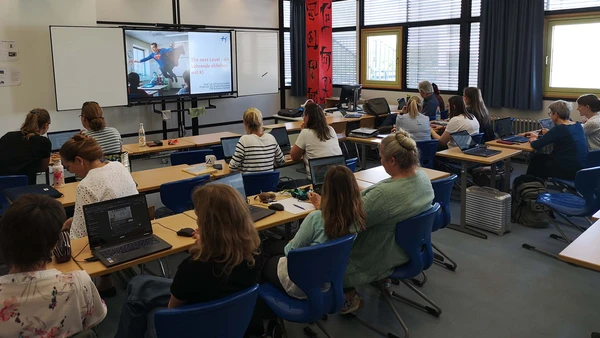 A lecture theatre from behind, you can see a large number of adults with laptops in front of them.