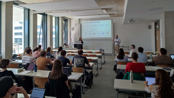 Ein Vorlesungssaal, vorne ein Mann, der etwas erzählt, und eine Frau am Computer, dazwischen an der Leinwand eine Präsentation, gegenüber eine große Anzahl Studierender.