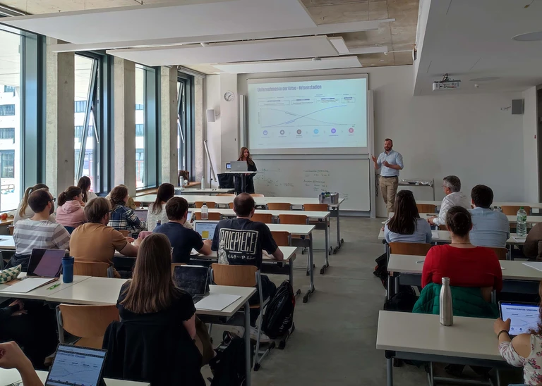 Ein Vorlesungssaal, vorne ein Mann, der etwas erzählt, und eine Frau am Computer, dazwischen an der Leinwand eine Präsentation, gegenüber eine große Anzahl Studierender.