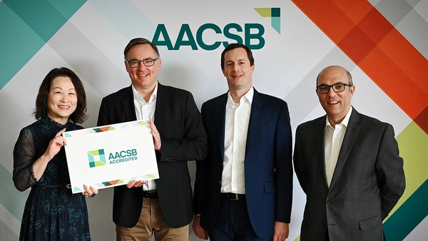 Vier Personen lächeln in die Kamera, zwei davon halten gemeinsam ein Schild in die Höhe auf welchem steht: „AACSB accredited“.