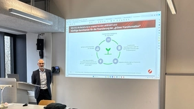 Ein Mann im Anzug steht in einem Hörsaal, neben ihm sieht man eine Präsentation an der Wand.