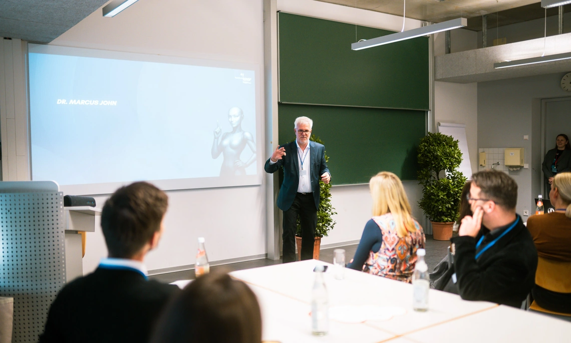 Männliche Person hält vor einer Gruppe, an Tischen sitzend, einen Vortrag