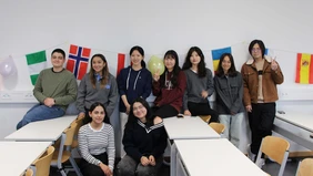 9 Personen stehen in einem Klassenzimmer und lächeln in die Kamera, im Hintergrund sieht man viele verschiedene Flaggen.