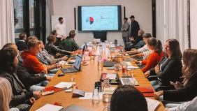 Eine Gruppe Menschen sitzt um einen großen Konferenztisch herum und verfolgt die Präsentation von drei Personen am Ende des Tisches. 